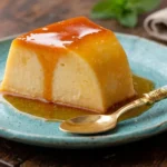 Fatia de pudim fit zero açúcar super cremoso com calda de caramelo escorrendo, servida em um prato azul rústico sobre uma mesa de madeira. Uma sobremesa saudável, fácil e sem leite condensado.